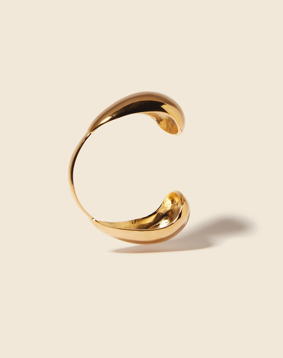 Bracelete ajustado ao pulso, com extremidades simétricas e curvas delicadas. Apresenta um acabamento banhado a ouro, adicionando um toque de sofisticação e brilho.