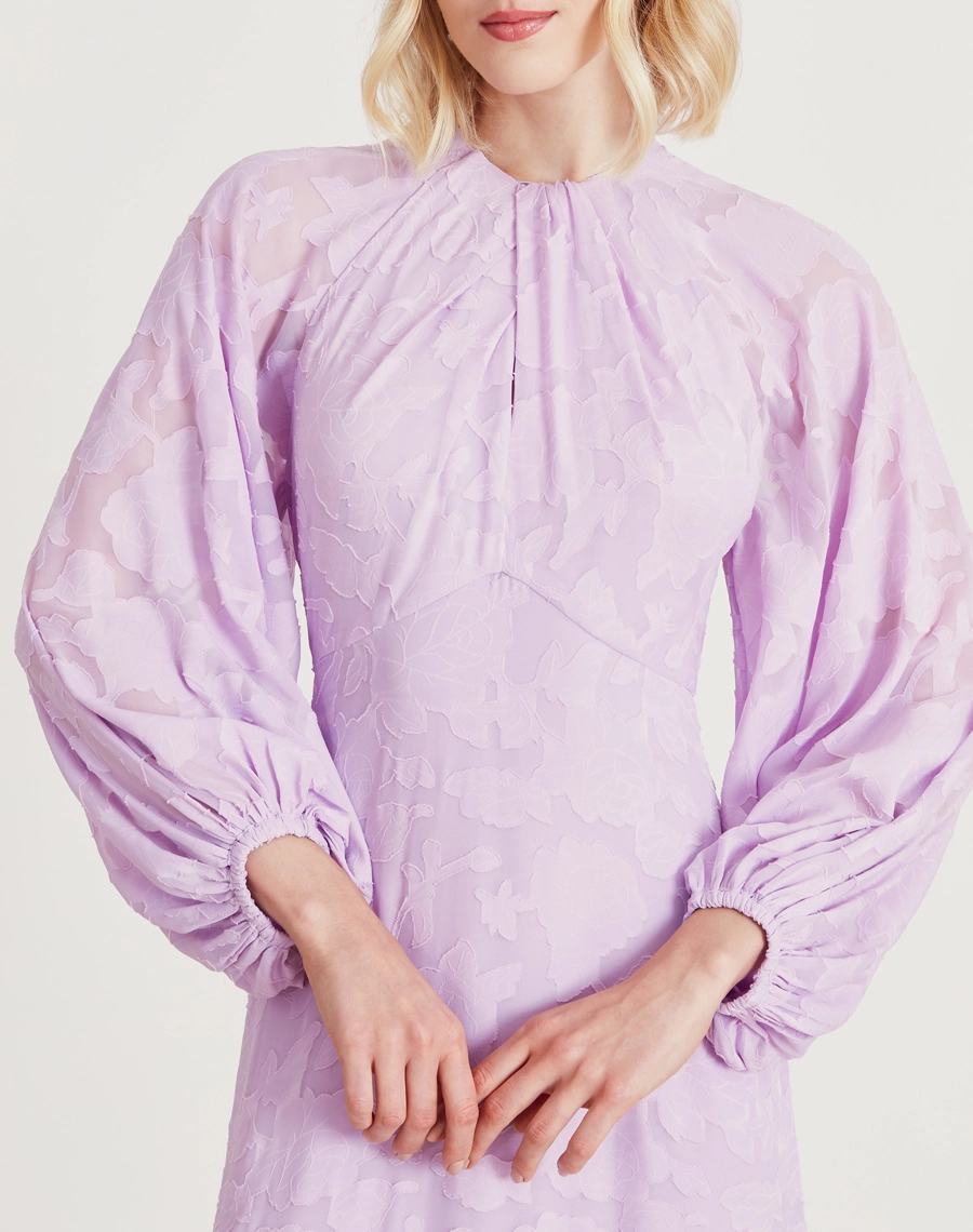 Vestido longo lavanda em jacquard floral com mangas bufantes e babado na barra. Sofisticação e elegância para eventos especiais.